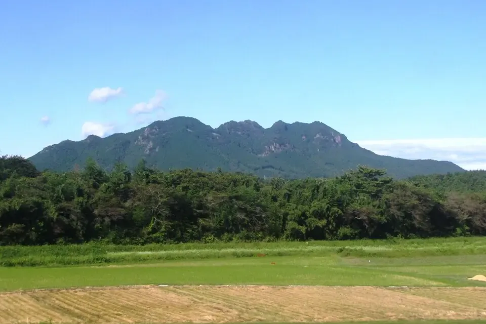 田畑と林の向こうに、ぎざぎざの稜線を持つ山が青空を背にそびえる風景